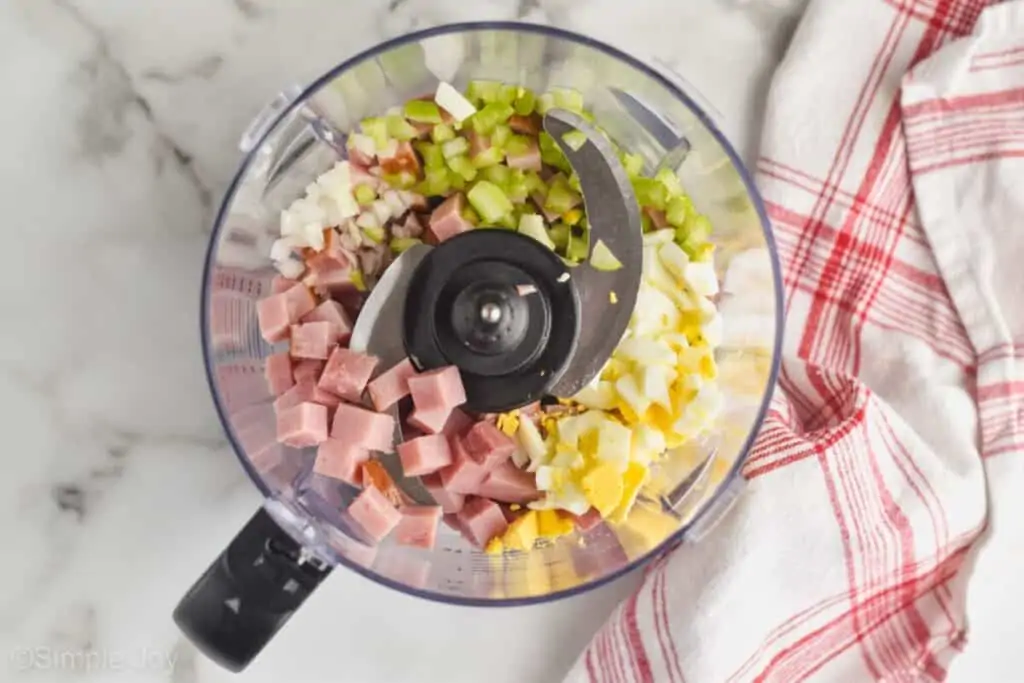 overhead view of a ninja food processor filled with ham, hard boiled eggs, celery, and onion to make ham salad