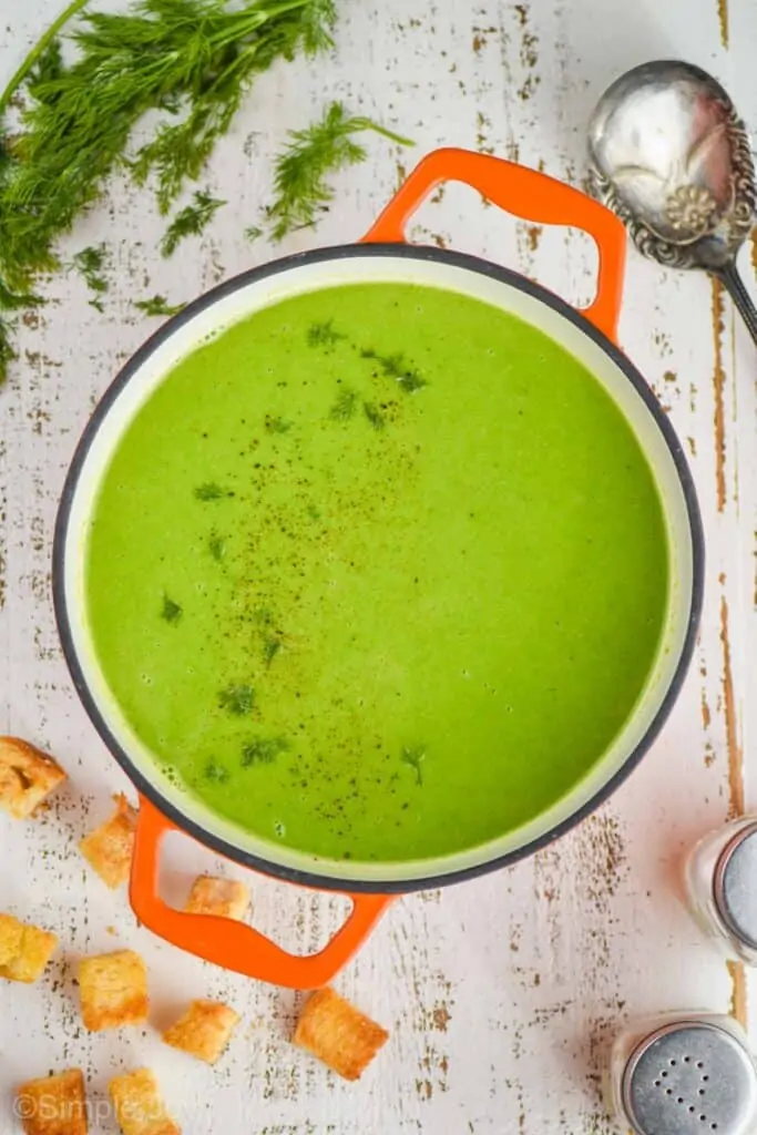 overhead view of an orange dutch oven filled with pea soup garnished with fresh dill on a white wooden surface