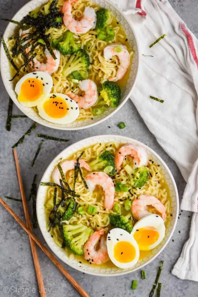 overhead view of two ramen bowls on a gray surface, each has shrimp, broccoli, and a soft boiled egg