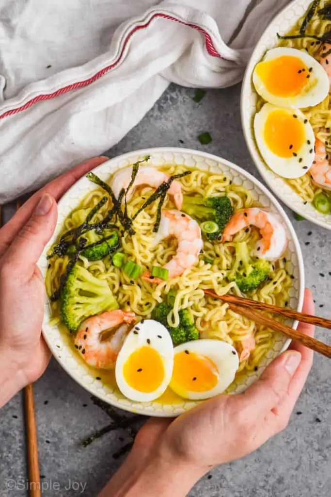 overhead view of two hands holding a bowl of easy ramen with a soft boiled egg, chopsticks, shrimp, and broccoli