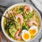 close up overhead view of a ramen recipe on a gray surface, bowl has small slices of seaweed over shrimp, broccoli, noodles, and soft boiled egg, all garnished with black sesame seeds - 7