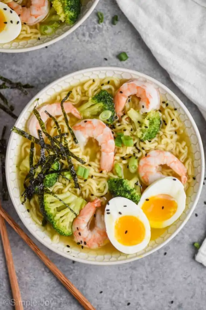 close up overhead view of a ramen recipe on a gray surface, bowl has small slices of seaweed over shrimp, broccoli, noodles, and soft boiled egg, all garnished with black sesame seeds