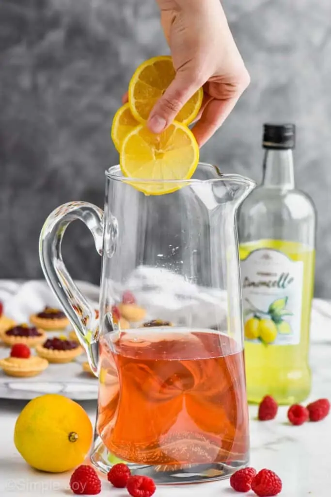 a hand dropping three lemon slices into a pitcher that has rose sangria in it