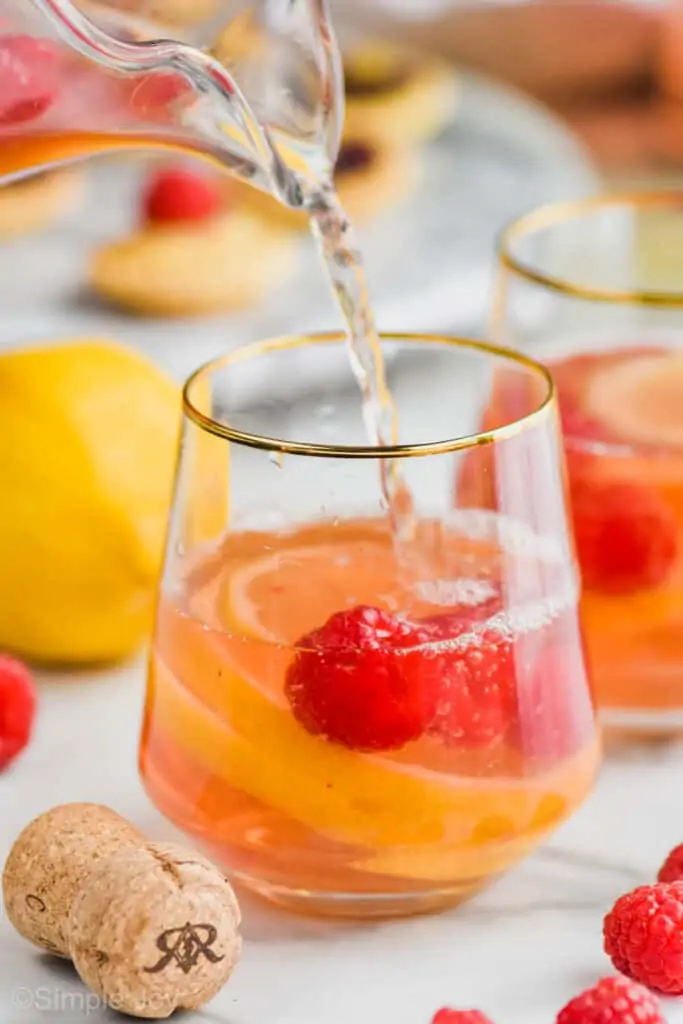pouring Sparkling Rosé Sangria into a gold rimmed stemless wine glass with raspberries and lemons in it