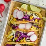 overhead of four vegan black bean tacos on a rimmed baking tray, garnished with sliced radishes, shredded cabbage, and corn