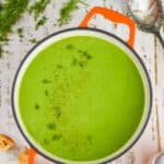 overhead view of an orange dutch oven filled with pea soup garnished with fresh dill on a white wooden surface