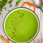 overhead view of an orange dutch oven filled with pea soup garnished with fresh dill on a white wooden surface