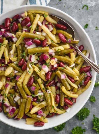 overhead of white bowl with three bean salad and a spoon in it on a gray surface garnished with parsley and on a cloth napkin
