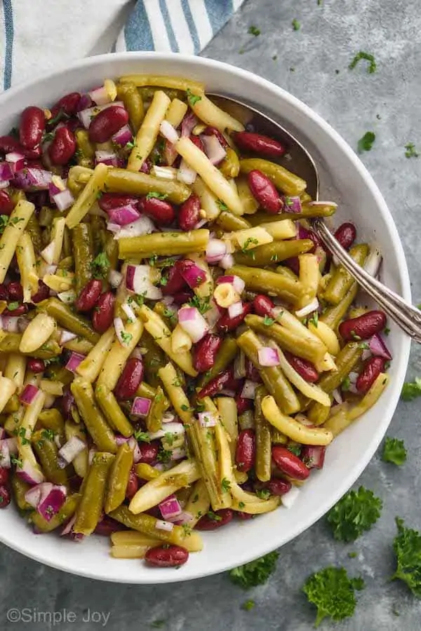 overhead of white bowl with three bean salad and a spoon in it on a gray surface garnished with parsley and on a cloth napkin