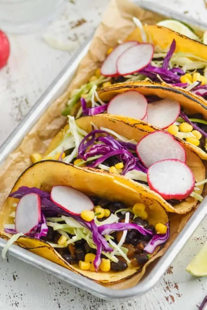 side view of four black bean tacos on a small baking tray lined with parchment paper, garnished with radishes and cabbage