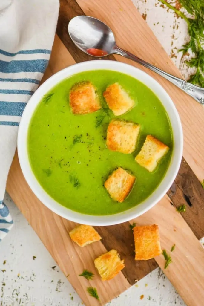 overhead view of a white bowl filled with pea soup recipe with five homemade croutons on a cutting board with a cloth napkin next to it