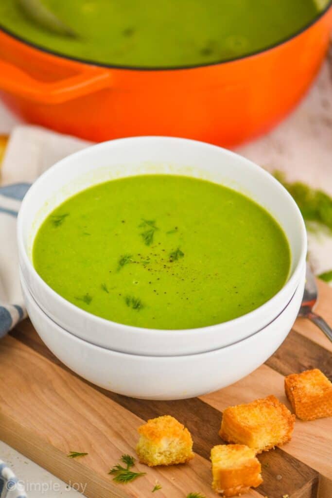 two white bowls stacked with pea soup in them on a cutting board with croutons around it and garnished with dill