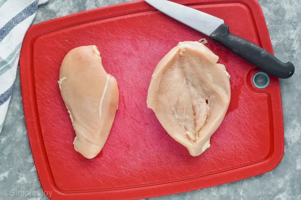 two raw chicken breasts on a red cutting board next to a knife with one being butterflied open