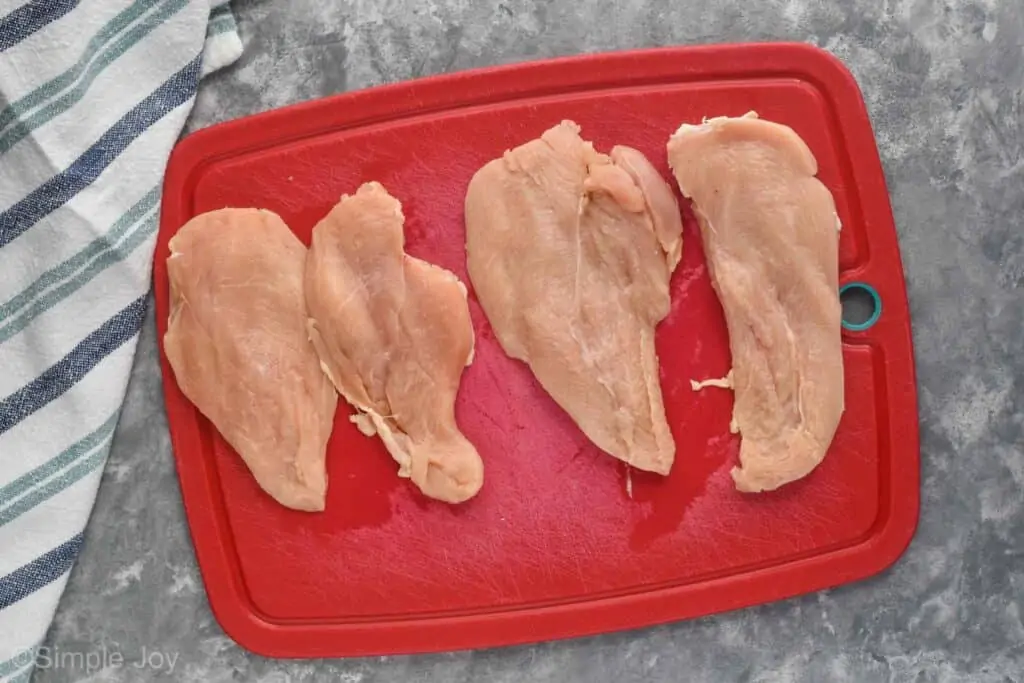 four chicken breasts (cut from two thick breasts) on a red cutting board next to a striped cloth napkin on a gray counter top