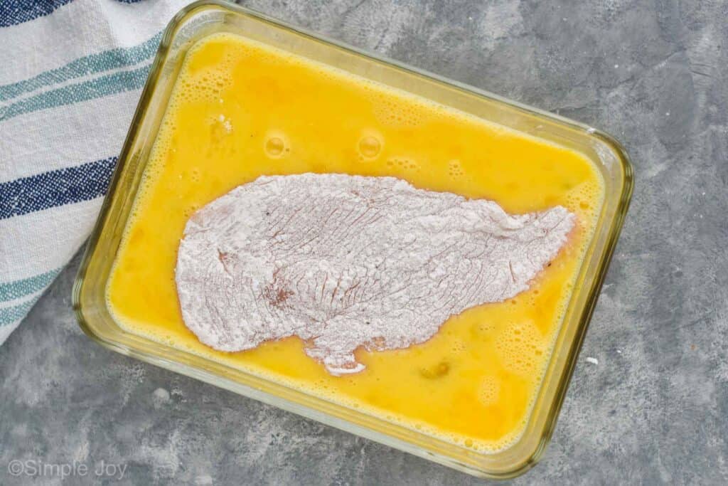 a rectangular baking dish with an egg mixture, and a floured chicken breast inside on top of a gray countertop