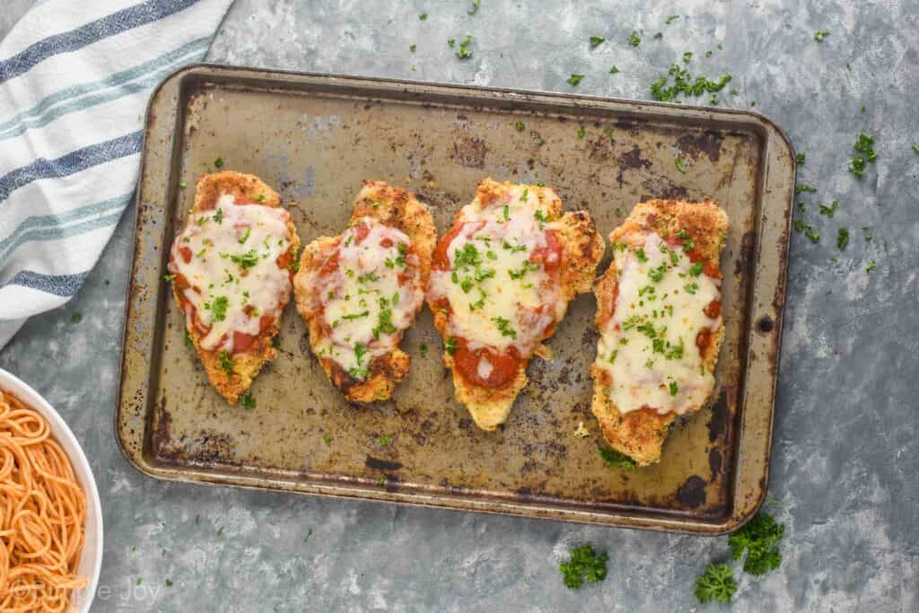 overhead view of four pieces of baked chicken parmesan on a vintage baking sheet, garnished with fresh parsley