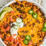 overhead view of a taco casserole in a white skillet with chopped tomatoes, jalapeños, sour cream, olives, and lime wedges