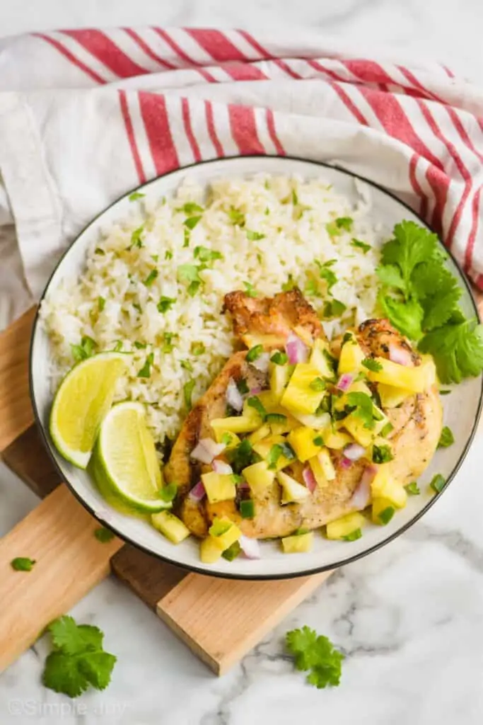 a grilled chicken breast covered in pineapple salsa on a plate with cilantro lime rice, lime wedges, and cilantro on a small cutting board with a striped cloth napkin