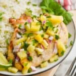close up side view of the best marinated grilled chicken breast that is covered in pineapple salsa with fresh cilantro next to it on a plate