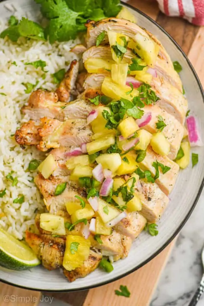 a close up of a sliced grilled chicken breast that was marinated covered in pineapple salsa on a black rimed white plate next to some rice