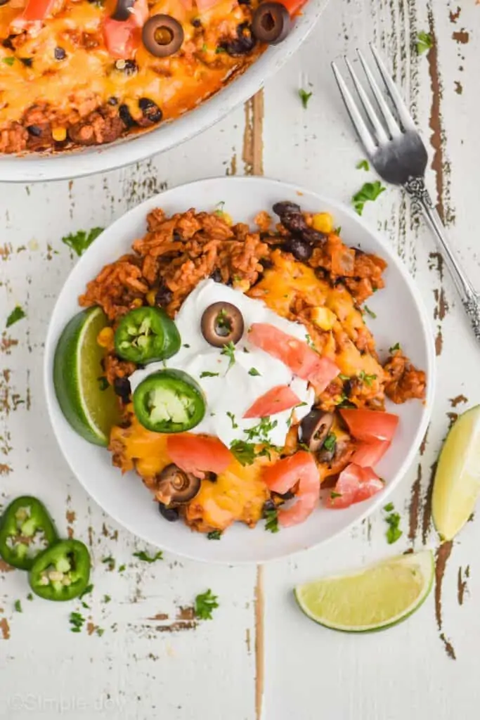 overhead view of a white plate with taco bake casserole on it, garnished with jalapeño slices, olive slices, diced tomatoes, sour cream, and chopped cilantro