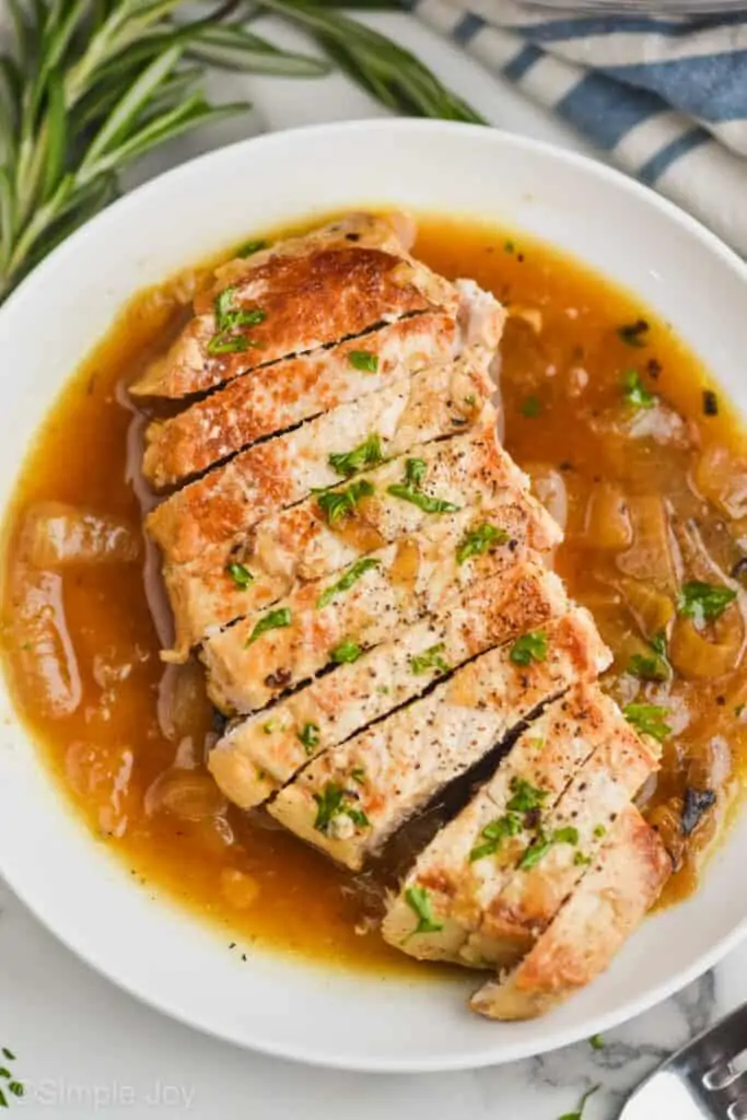 a chopped up boneless pork chop that has been pan fried, sliced, on a white plate with onion sauce around it, and covered with fresh parsley