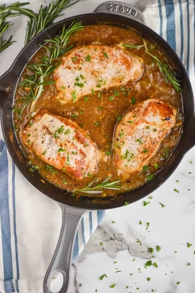 overhead view of a gray frying pan with three pork chops in it and a brown sauce, with fresh rosemary stems and chopped up parsley on a white marble countertop and a cloth napkin