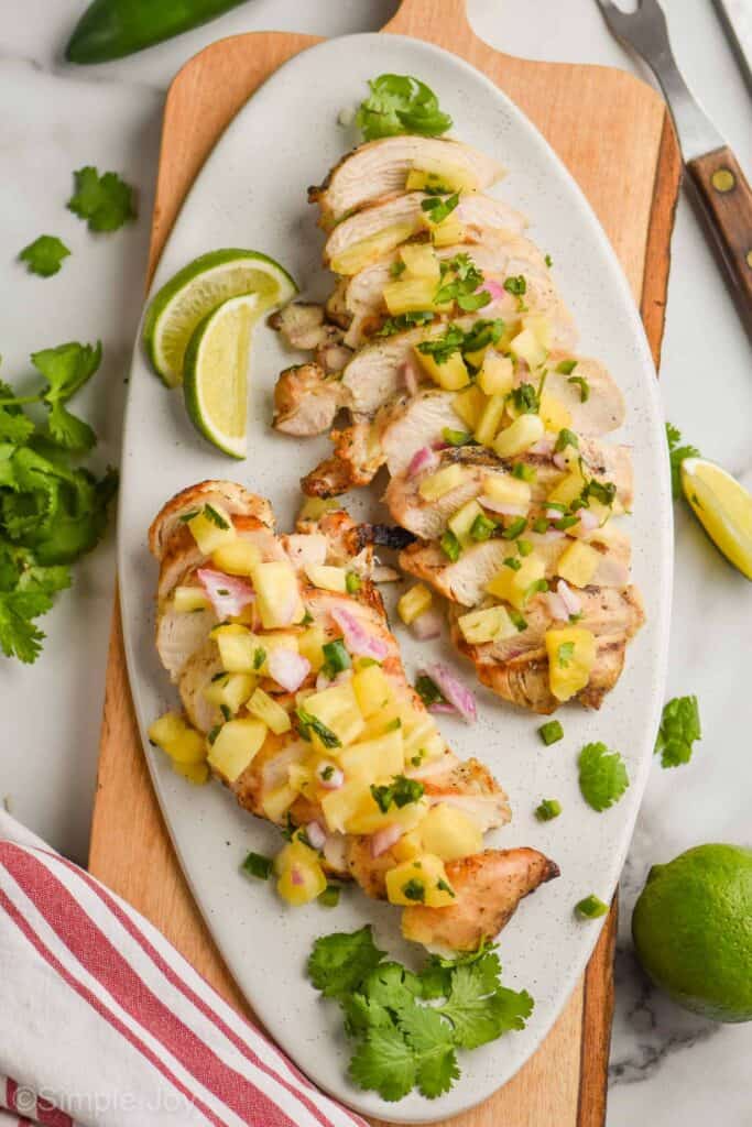 two grilled, sliced chicken breasts covered in pineapple salsa on a white oval plate on a large cutting board with lime wedges and fresh cilantro