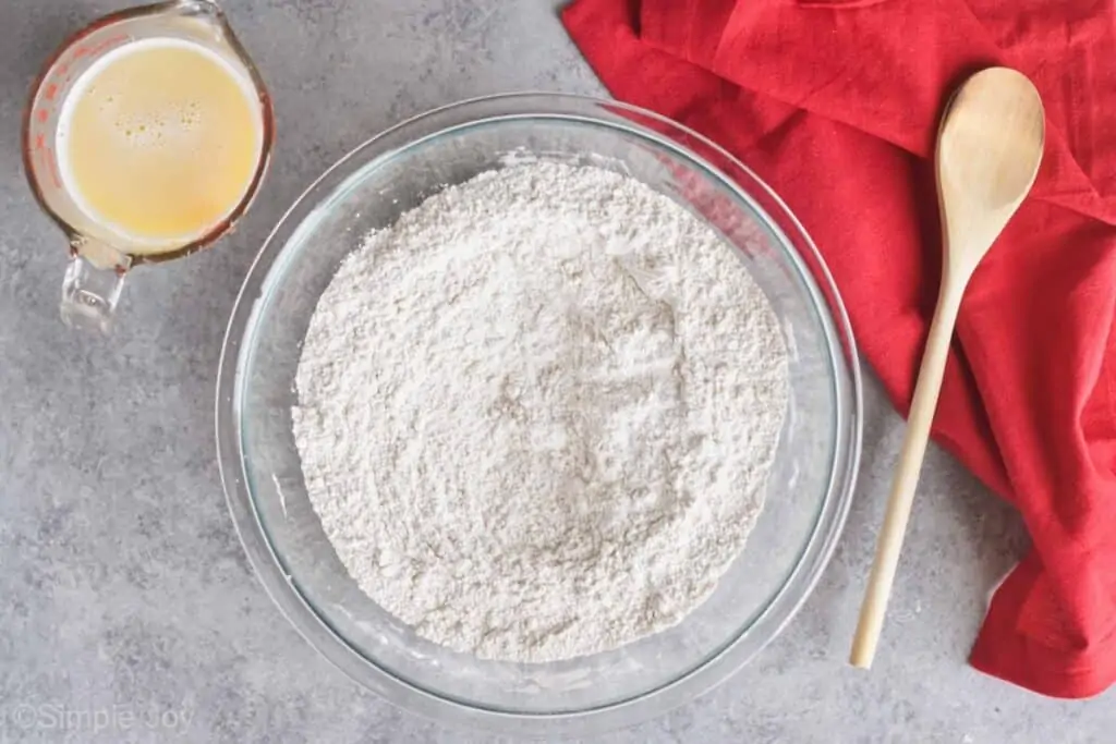 overhead view of a bowl of flour with a wooden spoon next to it and a glass measuring cup filled with melted butter and warm milk