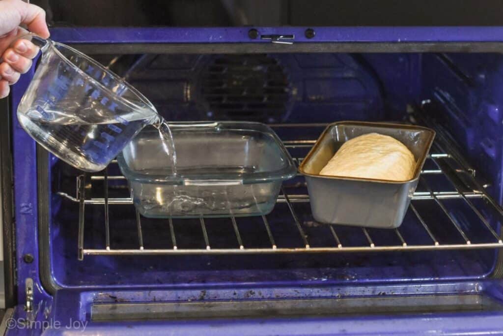 a loaf of sandwich bread dough in a bread pan in an oven with a hand pouring hot water into a square baking dish