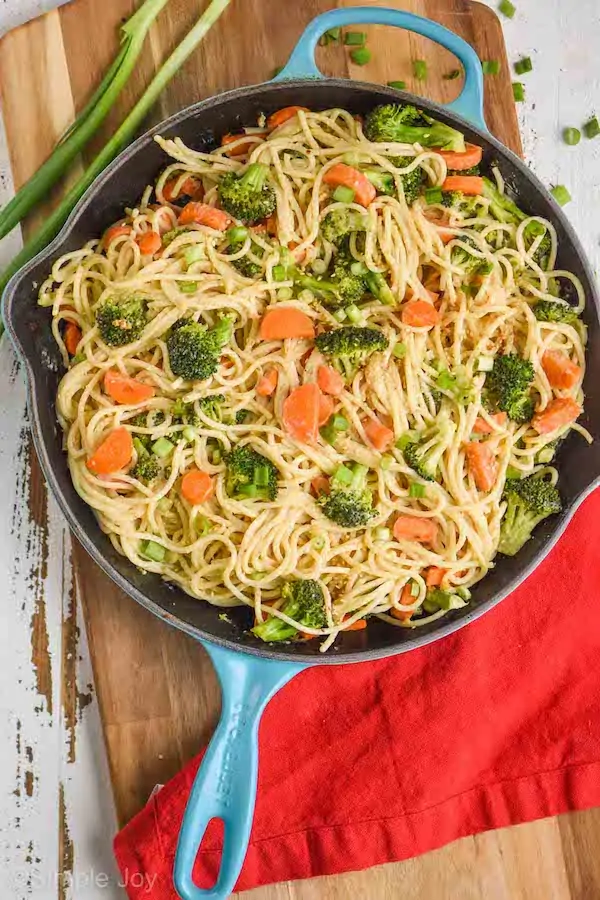 a cutting board with a teal skillet on it, inside is hummus pasta: spaghetti with sautéed carrots and broccoli that has a hummus based sauce