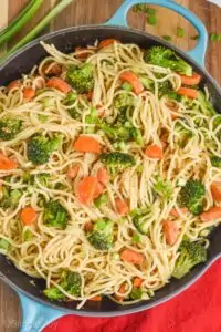 a close up of a teal skillet that has vegetables and spaghetti in it and is garnished with green onion slices and sesame seeds