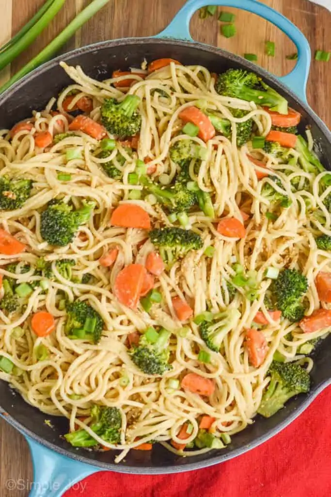 a close up of a teal skillet that has vegetables and spaghetti in it and is garnished with green onion slices and sesame seeds