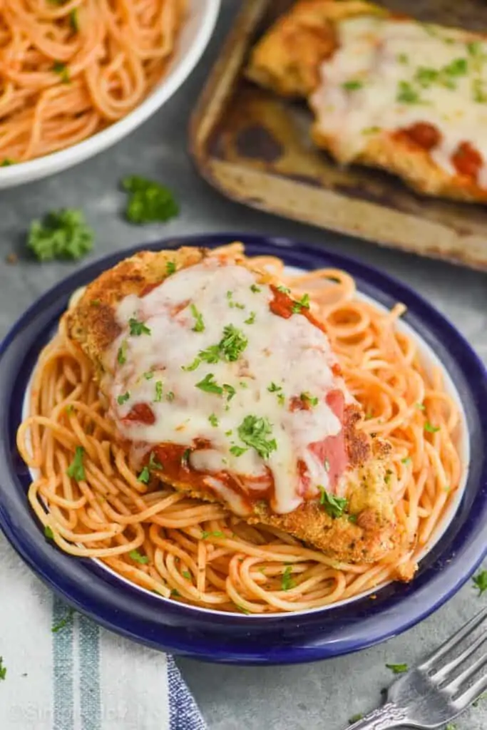 a piece of baked chicken parmesan that has been garnished with fresh parsley on a plate of spaghetti with a blue rim, with a tray of more chicken and a bowl of more spaghetti in the background