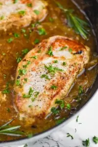 a pan fried pork chop in a a frying pan, with easy onion sauce around it and chopped parsley on top