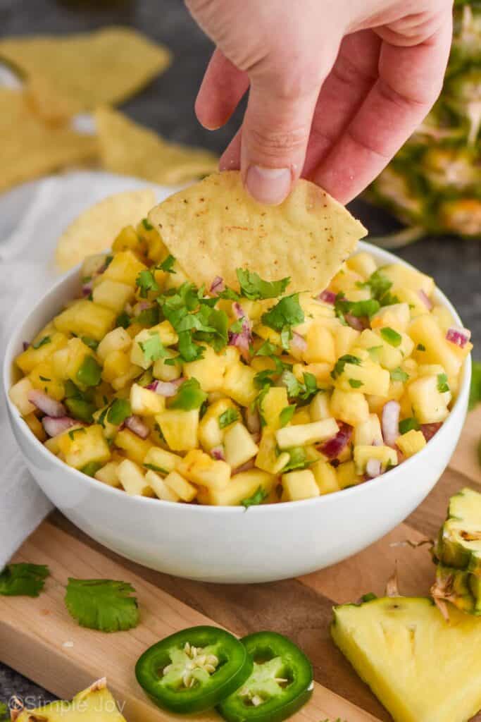 a white bowl full of pineapple jalapeño salsa on a cutting board with a hand dipping in a tortilla chip