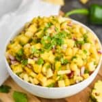 a white bowl with pineapple salsa in it, topped with fresh cilantro on a cutting board with pineapple chunks and jalapeños around it