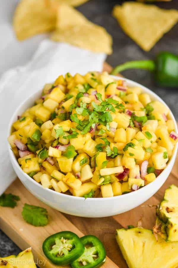 a white bowl with pineapple salsa in it, topped with fresh cilantro on a cutting board with pineapple chunks and jalapeños around it