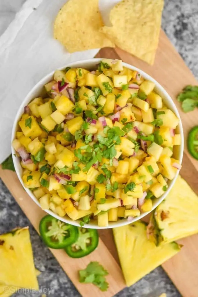 overhead view of a white bowl full of pineapple salsa, garnished with cilantro, on a cutting board with chunks of pineapple and jalapeño slices around