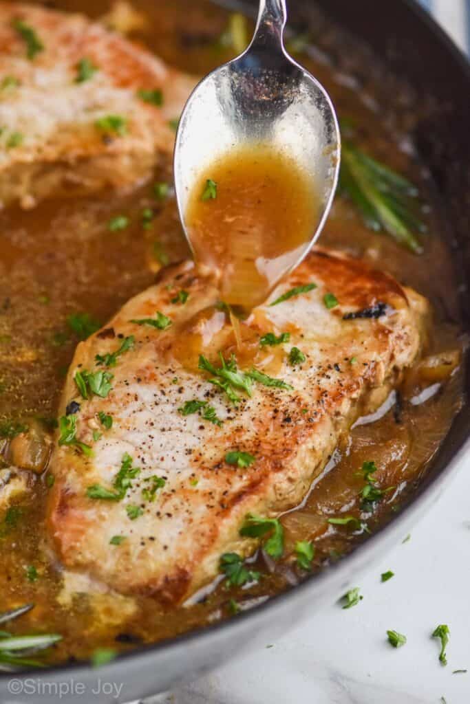 a spoon, spooning a brown sauce with onions onto a pan fried pork chop in a pan