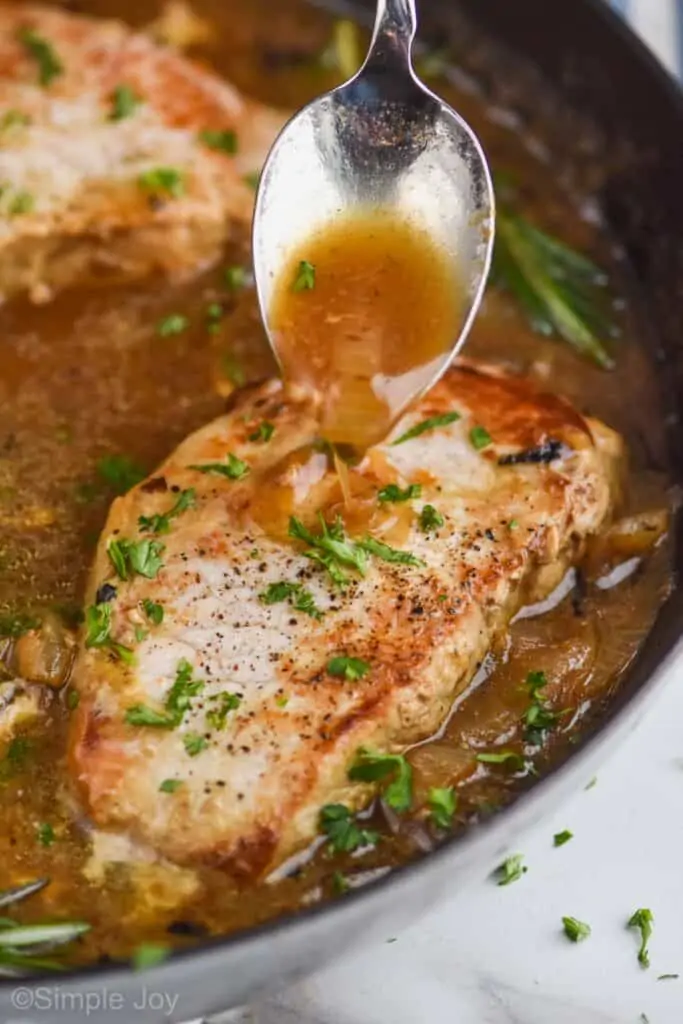 a spoon, spooning a brown sauce with onions onto a pan fried pork chop in a pan