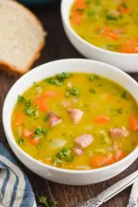 a white bowl on a wood surface full of split pea soup, garnished with fresh parsley