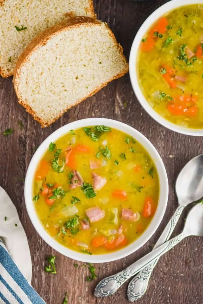 overhead view of a white bowl filled with spit pea and ham soup recipe with chunks of ham, carrots, and fresh parsley visible, two pieces of bread, two spoons on a wood surface