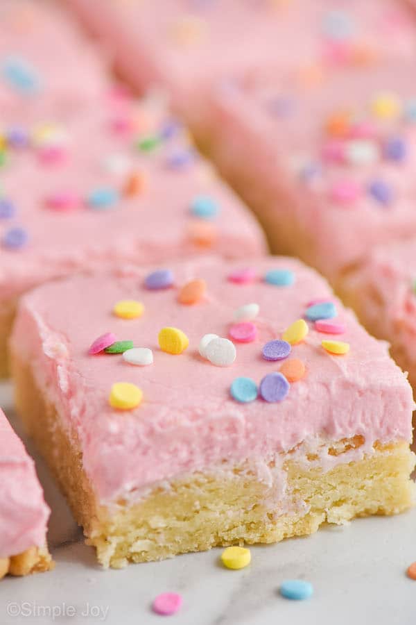 close up of a sugar cookie bar with pink buttercream frosting and circle pastel sprinkles on a white surface