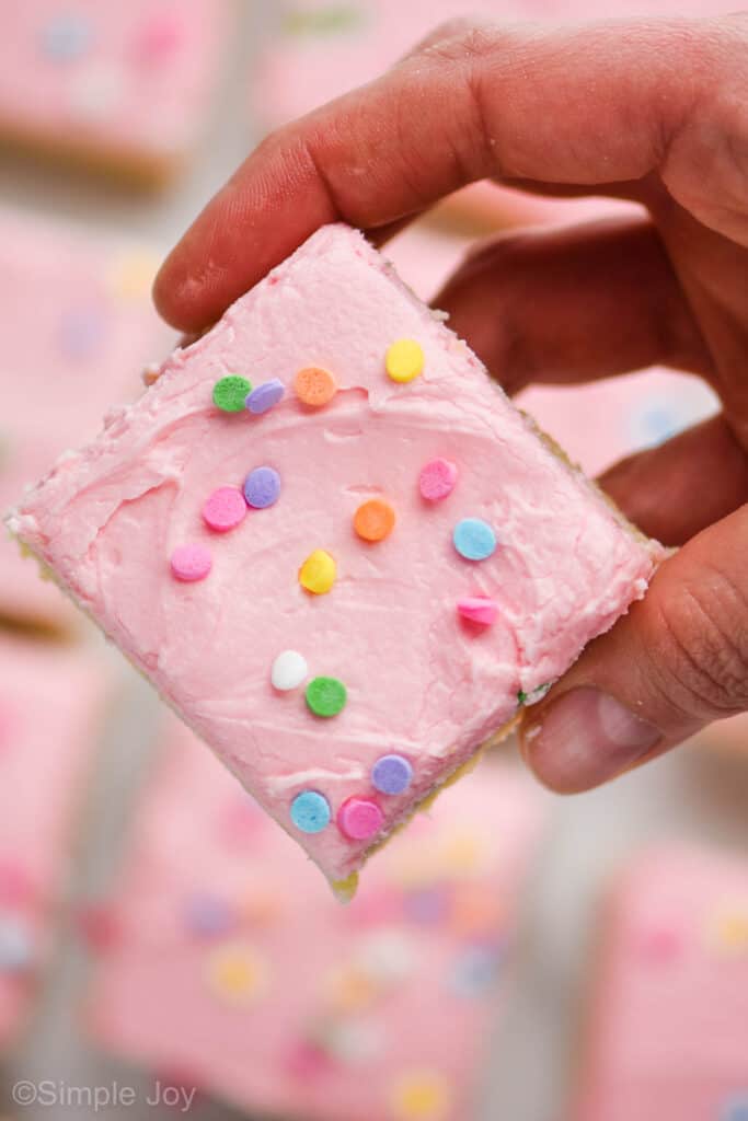 a hand holding up a square sugar cookie bar with pink frosting and sprinkles over other bars