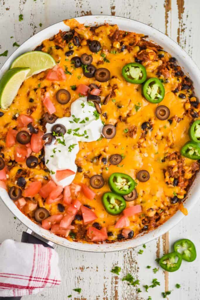 overhead view of a taco casserole in a white skillet with chopped tomatoes, jalapeños, sour cream, olives, and lime wedges