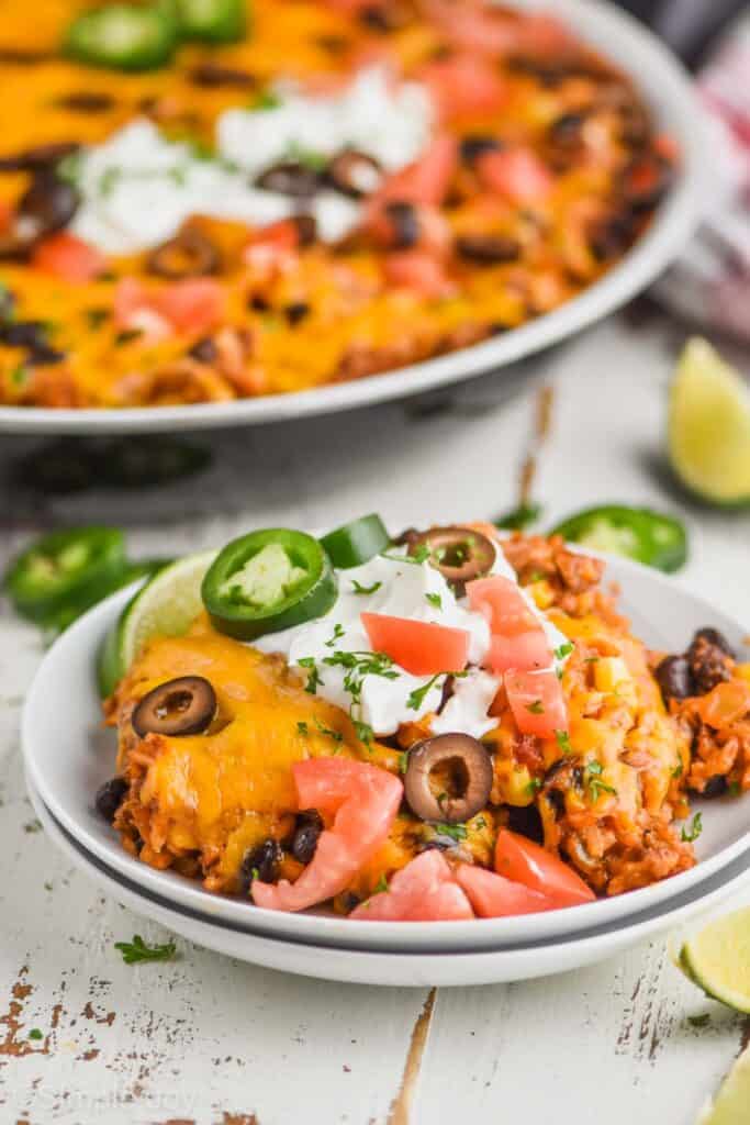 two small white plates stacked with a taco casserole recipe on them, garnished with olive slices, chopped tomatoes, and jalapeño slices, a skillet with taco casserole in the background