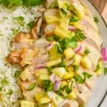a close up of a sliced grilled chicken breast that was marinated covered in pineapple salsa on a black rimed white plate next to some rice