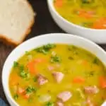 a white bowl on a wood surface full of split pea soup, garnished with fresh parsley