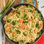 a cutting board with a teal skillet on it, inside is hummus pasta: spaghetti with sautéed carrots and broccoli that has a hummus based sauce